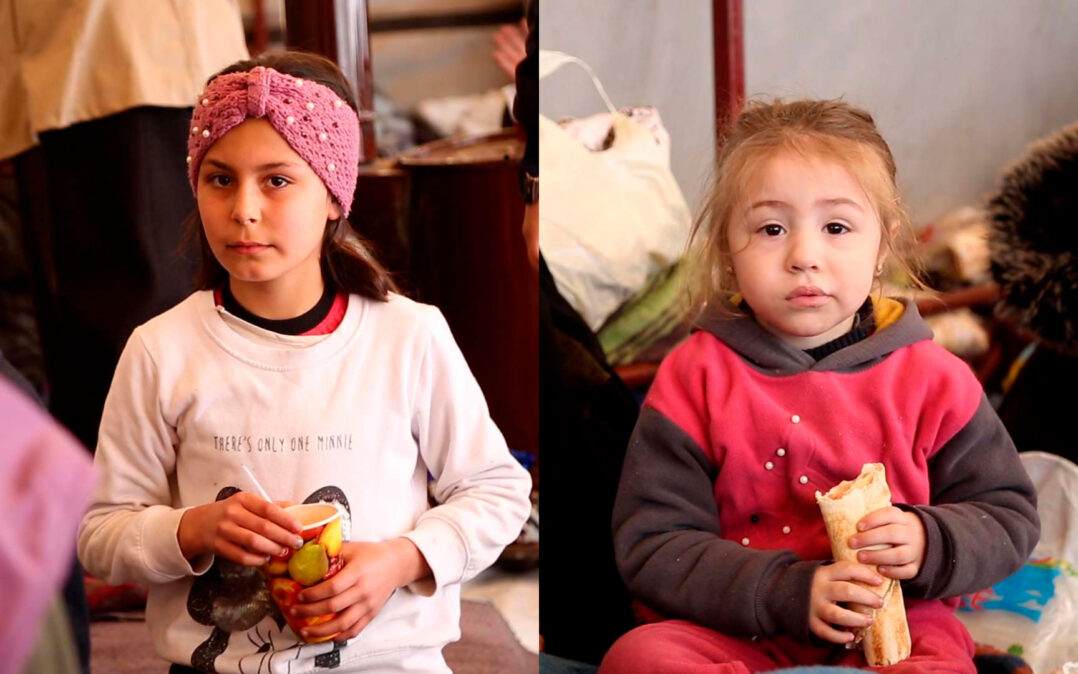 Kaksi lasta väliaikaisessa telttamajoituksessa, toisella kädessään paperimuki ja toisella leipä.
