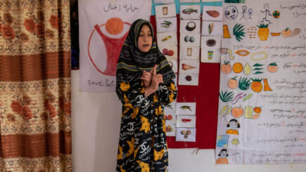 Nuori tyttö puhuu koululuokan edessä taustallaan erilaisia kuvallisia opetusmateriaaleja ja piirretty Pelastakaa Lapset -logo