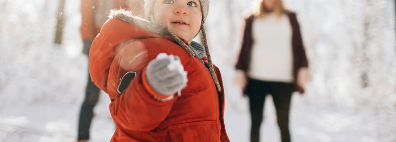 Pieni lapsi talvivaatteissa lunta kädessä, taustalla kaksi aikuista.