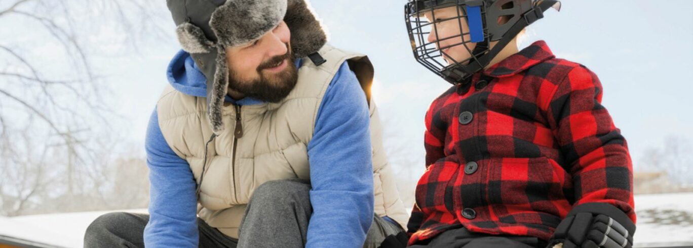 AIkuinen ja lapsi istuvat vierekkäin penkillä, lapsella päässä jääkiekkokypärä.