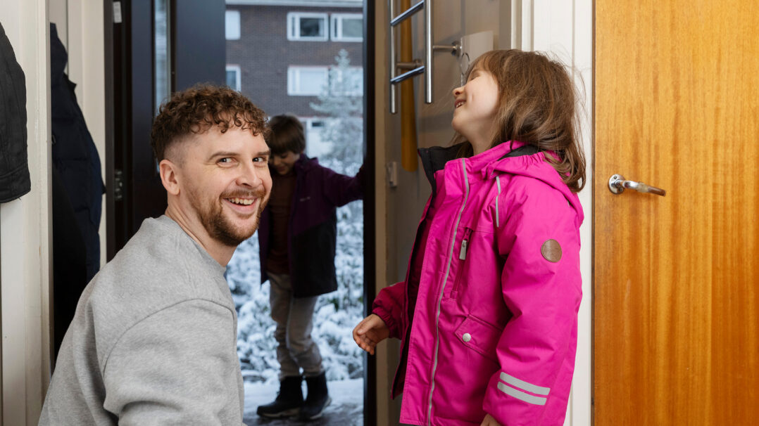 Mies ja lapset eteisessä. Mies katsoo hymyillen kameraan edessään hymyilevä lapsi. Taustalla poika odottaa ovensuussa takki ja kengät päällään.