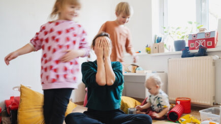 Kolme lasta leikkii taustalla. Nainen on lasten keskellä istumassa ja peittää käsillään kasvonsa.