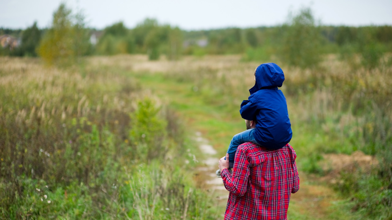 Lapsi istuu aikuisen harteilla ja katsoo maisemaa. Aikuinen ja lapsi ovat selin kameraan.